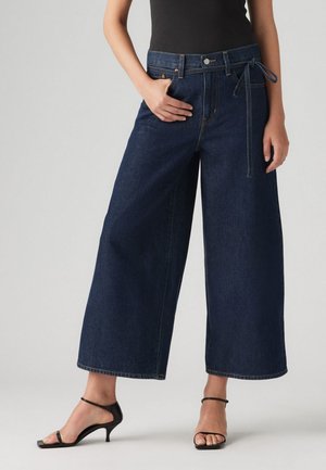 Dark blue wide-leg jeans made of denim, featuring a high waist, front pockets, and a belted tie detail. Paired with black strap heels.