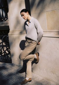 Sötét hajú nő kontyban, csíkos inget és bézs nadrágot visel, egy lábával a kőfalnak dőlve, amelyen a lába is nyugszik.