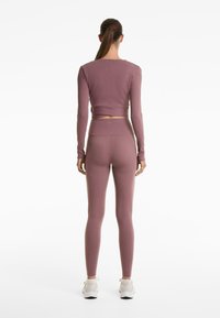 Žena stoji okrenuta leđima, nosi mauve dug rukav crop top i visoke tajice, a na nogama ima bijele tenisice, na bijeloj pozadini.
