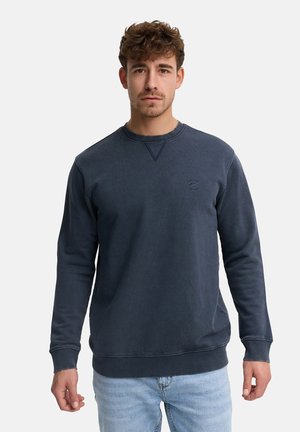 Man met kort krullend haar, gekleed in een donkerblauwe trui met ronde hals en lichtblauwe spijkerbroek, staand tegen een effen achtergrond.