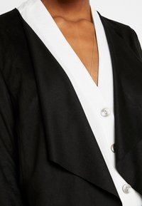 Sort yderlag med hvid krave og knapdetaljer. Blød tekstur med lagdelt design. Indeholder synligt stof, der draperer.