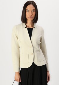 Blazer beige clair avec un col, deux poches avant et un ourlet arrondi. Le tissu a une texture subtile et est associé à un haut noir.