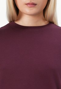 Burgundy t-shirt med rund halsringning, tillverkad av mjukt bomullstyg. Ytan är jämn med en lätt lyster och utan synliga mönster eller dekorationer.