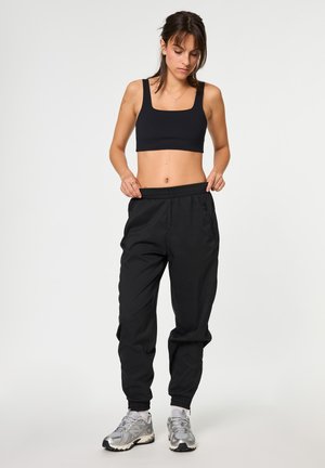 Kvinde iført sort crop top, sorte joggingbukser og sølv sneakers, justerer linningen, står på ensartet lyst baggrund.