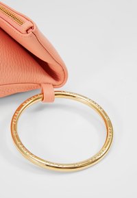 Persikofärgad läderclutch med guldringhandtag. Väskan har en texturerad yta, dragkedjestängning och ingraverade detaljer på ringen.