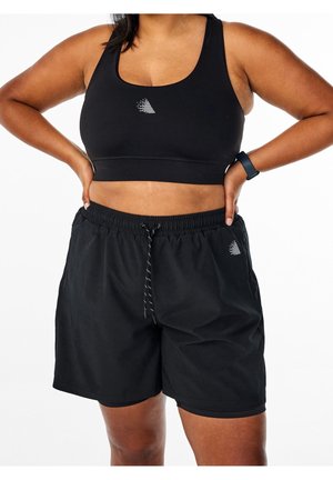Femme portant un soutien-gorge de sport noir et un short noir à cordon, les mains sur les hanches, montrant le ventre et un tracker d'activité au poignet.