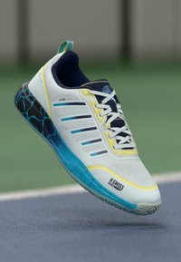 Sapatilha atlética branca com detalhes azuis, parte superior texturizada e solado estampado. Apresenta realces amarelos e seis ilhós para os atacadores.