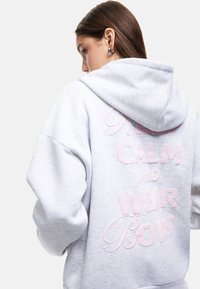 Grå hoodie med en stor lyserød broderet slogan på ryggen. Har hætte og løs pasform, lavet af blødt, tekstureret materiale.