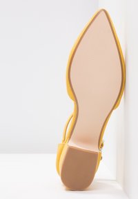 Chaussure à talon haute en suède jaune avec un bout pointu, dotée d'une semelle lisse beige à texture nervurée et d'un détail de fine sangle.