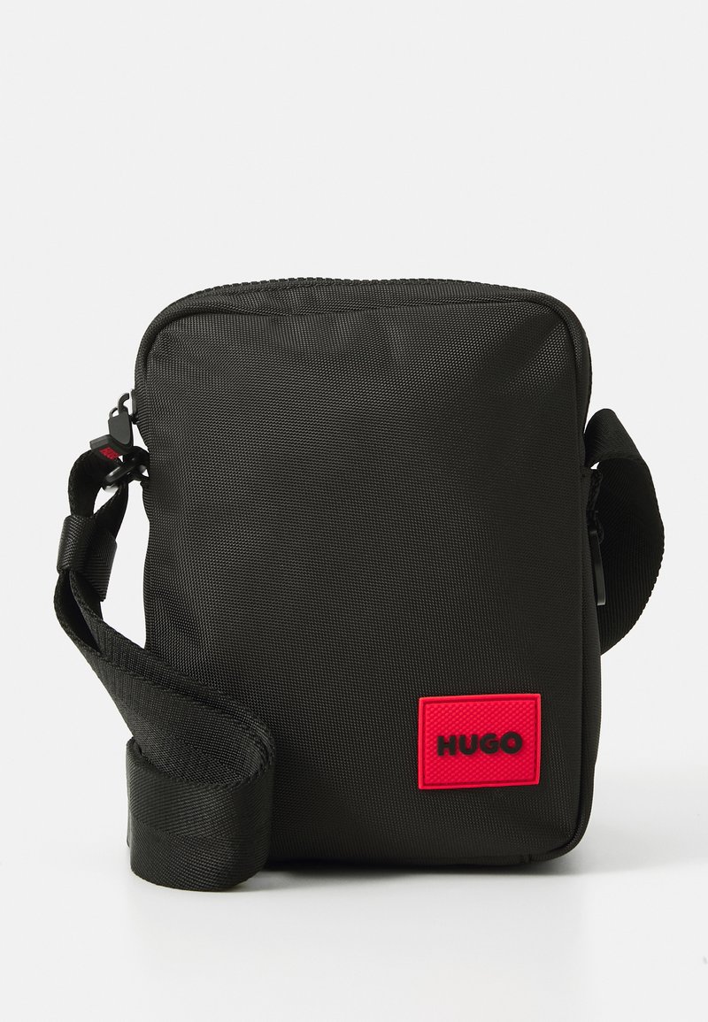 La borsa a tracolla nera in nylon presenta una chiusura con zip, una tracolla regolabile e un grande patch del logo rosso HUGO sul fronte.