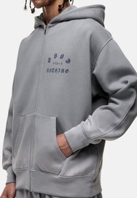 Felpa grigia con zip, tasche frontali, polsini a costine e una stampa grafica che recita "Dio per Niente" in blu scuro sul petto. Tessuto morbido.