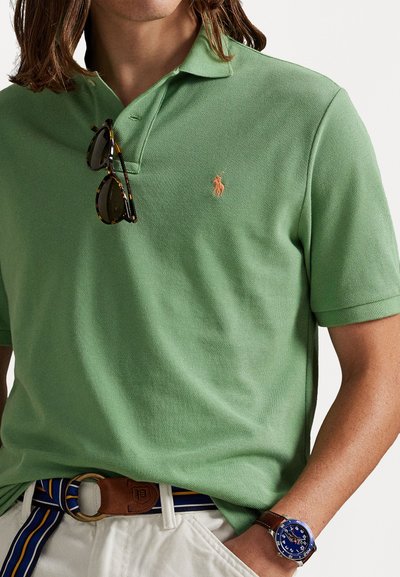 Zaļš polo krekls ar īsām piedurknēm un faktūru. Tiek piedāvāts ar izšūtu logotipu, kopā ar brūnu jostu un zilu pulksteni.