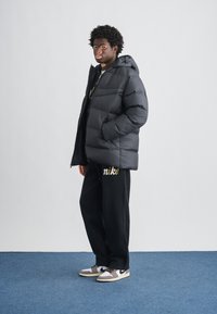 Czarna puchowa kurtka z kapturem, o fakturowanym wykończeniu, noszona na ciemnej bluzie z kapturem. W połączeniu z czarnymi spodniami dresowymi z złotym logo i sneakersami.
