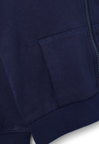 Sudadera azul marino con cremallera y bolsillo canguro, puños y dobladillo acanalados; textura suave con un acabado liso y detalles sutiles de costura.