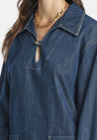 Femme portant une chaîne en or et une chemise en jean bleue avec un large col, une ouverture en forme de trou clé à l'avant et une fermeture par boutons.
