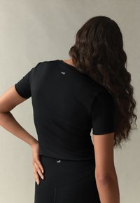 Svart kortärmad topp i stretchtyg med figurnära passform. Liten vit logotyptext placerad på ryggen nära halsringningen.