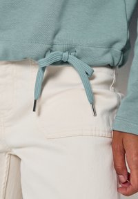 Gros plan d'un cordon de serrage bleu clair noué à l'ourlet d'un sweat-shirt assorti, associé à un pantalon blanc cassé et une main droite au repos.
