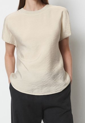 Vrouw die een beige, gestructureerd short-sleeve blouse draagt, ingestopt in een zwarte broek met handen in de zakken tegen een effen achtergrond.