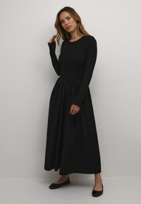 Musta pitkähiha dressi, jossa on istuva ribattu vartaloosa ja virtaava, nyöröity hame, valmistettu pehmeästä kankaasta. Litteät mustat kengät viimeistelevät ilmeen.