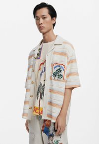 Korte mouwen, gestreept shirt in beige en lichtoranje met gouden knopen, voorzien van een geborduurd zakje met palmbomen en "BUENA VIDA."