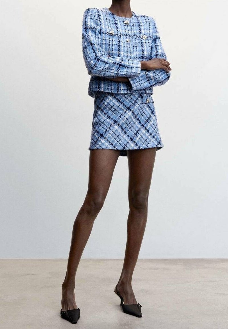 Ensemble blazer et jupe en tweed bleu avec un motif à carreaux, doté de boutons dorés et d'une silhouette ajustée. Accompagné de chaussures pointues noires.