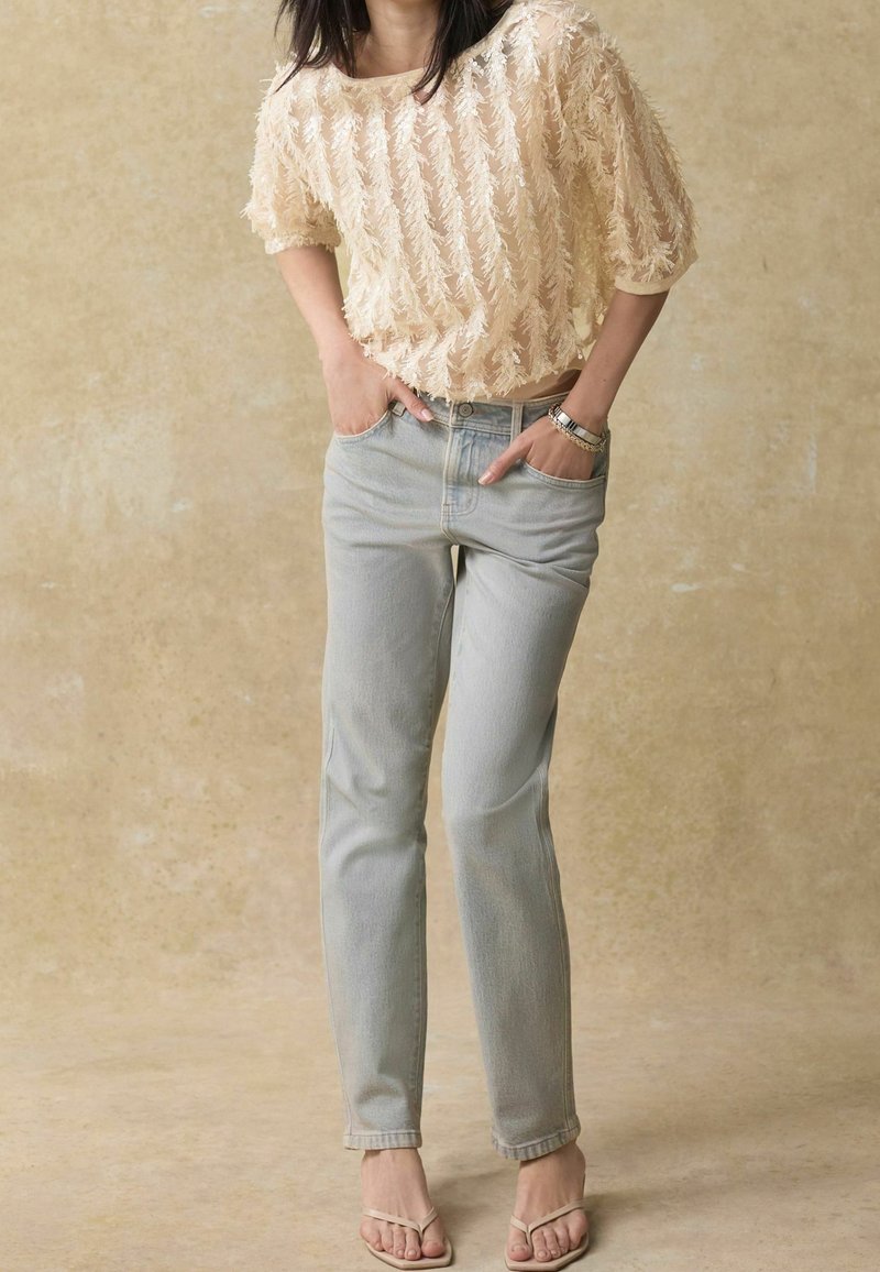 Blouse beige texturée en tissu transparent avec un motif de feuilles, associée à un jean droit bleu clair et des tongs minimalistes.