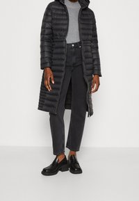 Sort puffet jakke med lang kant og hætte, prydet med quiltet tekstur og metalliske lynlåse, parret med en grå top og sorte jeans.