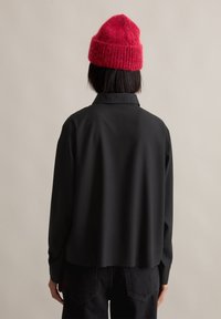 Svart skjorte med krage og lange ermer, med en glatt tekstur og lett buet hem, sammen med en rød strikket lue.