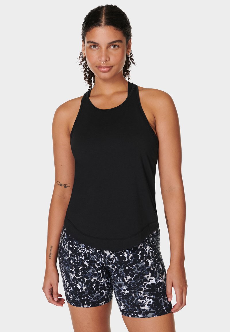 Vrouw met krullend haar, gekleed in een zwart mouwloos shirt en zwart, wit en grijs gestreepte shorts, staand tegen een lichte achtergrond.