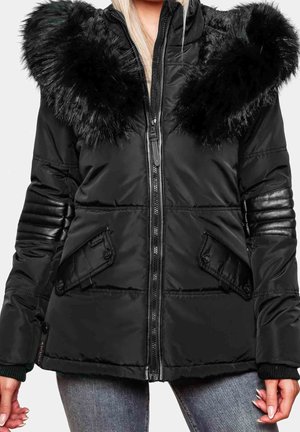 Chaqueta de invierno acolchada negra con gran cuello de piel sintética, cremallera frontal, parches acolchados en los codos y bolsillos con solapa y botones, usada con vaqueros azules.