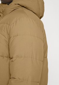 Sandfarvet pufferjakke med hætte, der har quiltede sektioner og en glat, mat tekstur. Subtil syning detaljer synlige på stoffet.
