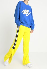 Blå sweatshirt med en vit Nike-logotyp, tillsammans med ljusgula byxor med svarta sidor och tryckknappar vid fållen.