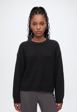 Giovane donna con lunghi capelli ricci intrecciati che indossa orecchini a cerchio dorati, un maglione nero strutturato e pantaloni grigi su sfondo bianco.