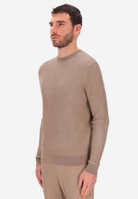 Uomo con capelli corti e scuri e barba, che indossa un maglione a coste taupe e pantaloni beige, in piedi davanti a uno sfondo bianco semplice.