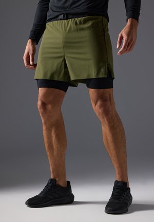 Olive green athletic shorts feature a waistband with a buckle, paired with black compression shorts. Material appears lightweight with side slits.