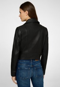 Svart läderjacka med kort croppad design, sned krage, slät yta och åtsittande ärmar, kombinerad med blå denimjeans.