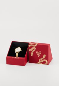 Gold watch with a white face, round casing, and thin strap inside a red box featuring snake design and logo. Black velvet lining visible.