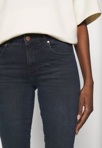 Mörkblå denimjeans med slim fit, med kopparknappar och subtila sömdetaljer, i kombination med en krämfärgad topp.