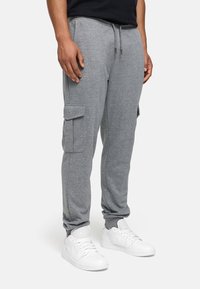 Pantalons de survêtement gris avec taille élastique et cordon de serrage ajustable, dotés de poches cargo latérales et de poignets fuselés. Portés avec des baskets blanches.