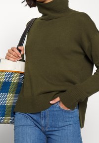 Maglione a collo alto a coste verde oliva con fessure laterali, abbinato a jeans azzurri e a una borsa a tracolla con motivo intrecciato nei colori blu, verde e bianco.