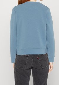 Felpa blu in cotone con maniche lunghe e polsini e orlo a costine, abbinata a jeans in denim scuro che mostrano dettagli delle tasche posteriori.