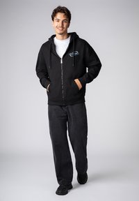 Black zip-up hoodie with a white interior, featuring a graphic logo on the left chest. Paired with loose black jeans and black shoes.