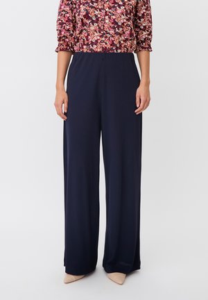 Donna che indossa pantaloni larghi blu navy, una maglietta floreale a manica lunga e tacchi beige con punta, in piedi su uno sfondo neutro.
