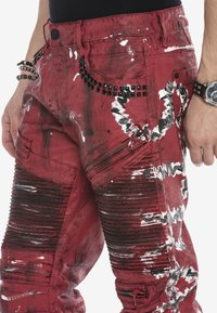 Rote Denim-Cargo-Hosen mit weißen und schwarzen Farbspritzern, Nietenakzenten und Reißverschlusstaschen. Mit gerippter Textur entlang der Beine.