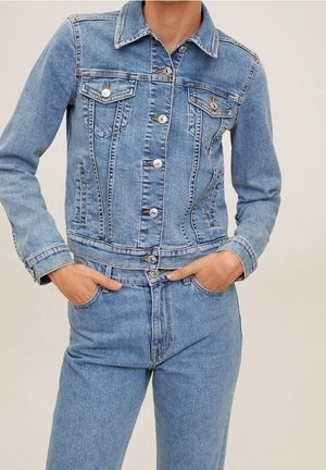 Personne portant une veste en jean bleu clair avec des boutons et un jean taille haute assorti, les mains dans les poches avant.