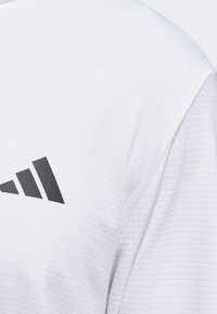 Valkoinen urheilupaita, jossa on teksturoitu kuvio ja vasemmalla puolella musta Adidas-logo. Kangas näyttää kevyeltä ja hengittävältä.