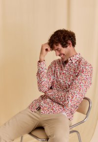 Overhemd met knoopsluiting in een rood en groen bladpatroon op een witte achtergrond; katoenen stof, lange mouwen, getailleerde pasvorm. Beige broek, metalen stoel.