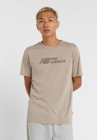 Camiseta de algodón beige con el logo de "new balance" en marrón oscuro. Manga corta, cuello redondo, textura suave, diseño minimalista.