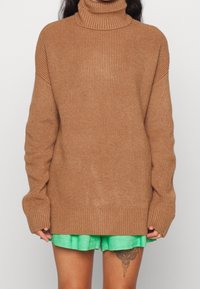 Brun ribbad polotröja med lång ärm och lös passform, i kombination med gröna shorts, som visar upp stickad textur och avslappnad design.