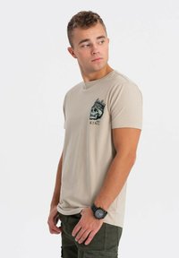 Béžové bavlněné tričko s černou grafikou lebky s korunou a textem "KRÁL," v kombinaci s olivově zelenými cargo kalhotami a hodinkami.